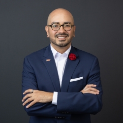 Headshot of Associate Vice President for Student Life and Dean of Students. Man is posing with his arms crossed; he is dressed in  a navy suit, adorned with a red flower like pin and a Puerto Rican Flag lapel pin. 